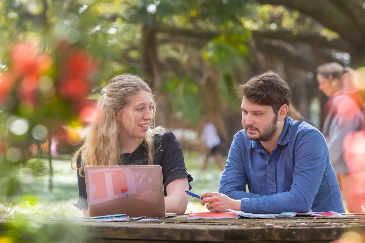 Two students sit at an outdoor table looking at a computer.
